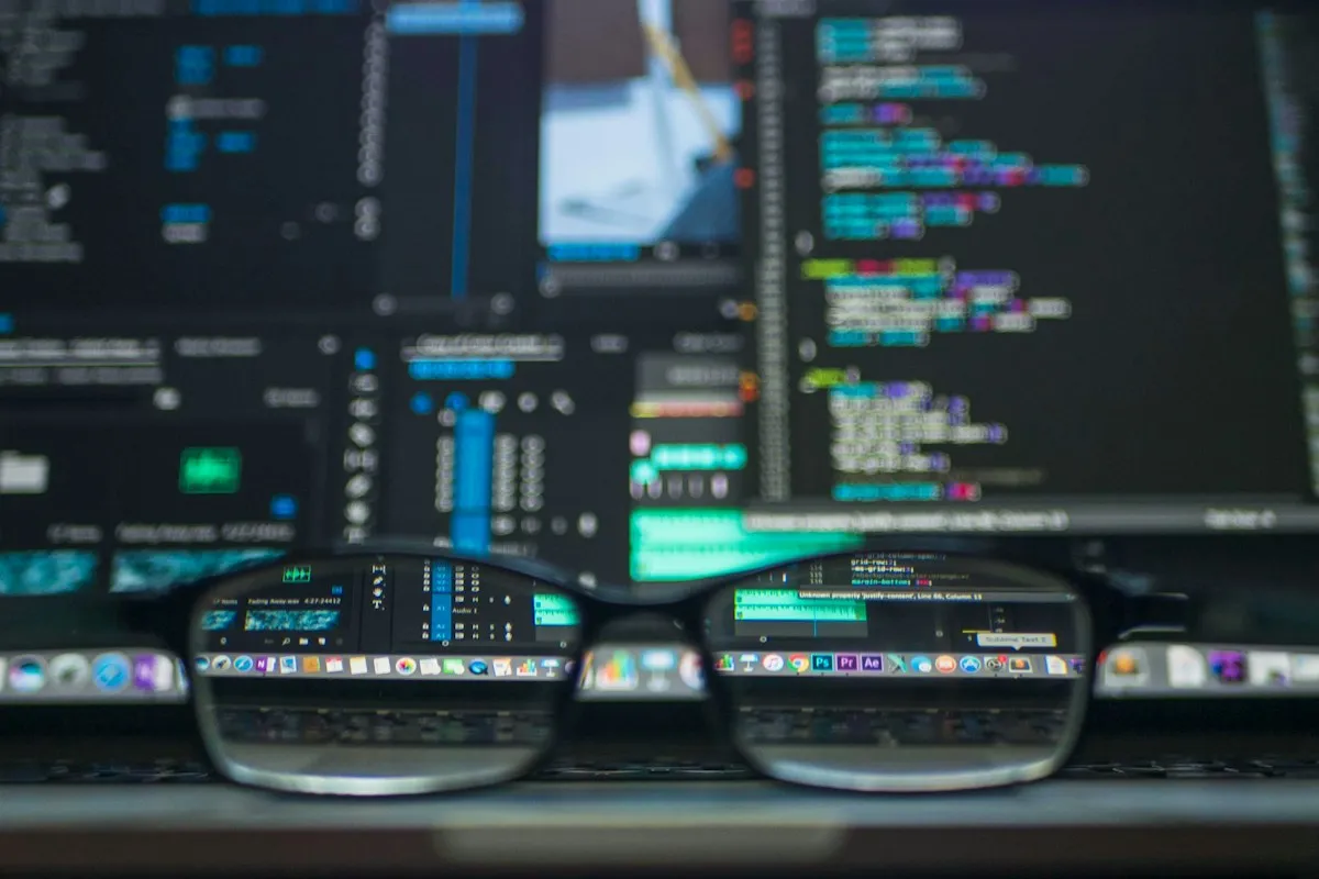 Brille vor zwei unscharfen Monitoren mit Quellcode und Schnittsoftware